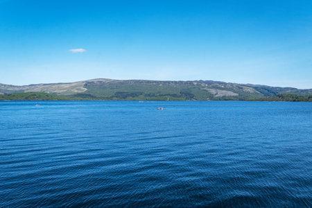 A wide, tranquil view across the calm blue waters of Loch Lomond from a gravel and pebble shore, with tree-covered hills under a bright blue sky.の写真素材