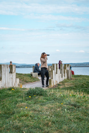 A person uses binoculars to look out over the sea from a coastal viewpoint on a partly cloudy day, with others sitting nearby.の写真素材
