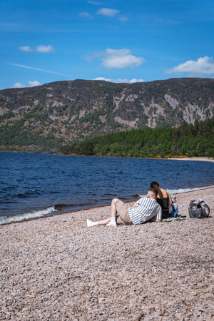 Two people relax on the scenic pebble beach at Dores, on the shore of Loch Ness, Scotland, enjoying the sunny day and tranquil views across the water.の写真素材