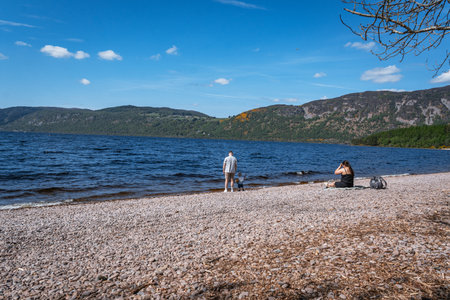 Two people relax on the scenic pebble beach at Dores, on the shore of Loch Ness, Scotland, enjoying the sunny day and tranquil views across the water.の写真素材