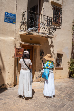 Two women in long skirts and hats stand on a cobbled street, looking at a traditional building with a balcony in a sun-drenched Greek town.の写真素材