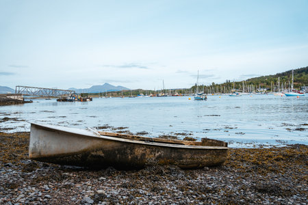 An old, weathered rowing boat rests on a shore covered in rocks and seaweed, facing a marina filled...