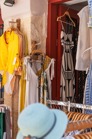 A clothing boutique displays women's summer apparel on racks in a sun-drenched alley, with a red door and a straw hat.の写真素材