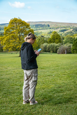 A woman stands in a large green field in a scenic landscape, looking at her phone with rolling hills...