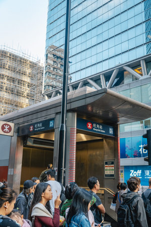 Commuters and tourists at the iconic MTR station entrance surrounded by modern glass skyscrapers.
