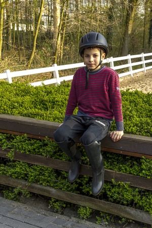 cute teenage  boy in helmet and boots for horse riding sitting next to manegeの写真素材
