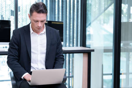 At a company a businessman is working on his laptop, laying it on his legs. He is preparing a meeting on the company's financial system. which has been assigned by the managementの写真素材