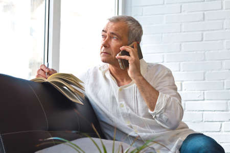 mature man with book and mobile phone on the sofa at homeの写真素材