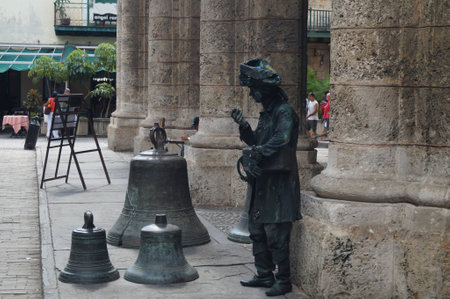In an enchanting plaza under the fading light, a street performer, dressed as a statue, interacts quietly with oversized bronze bells. Visitors admire the unique display, captivated by the artistry.の写真素材
