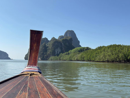 A longtail boat glides silently over calm waters, revealing towering limestone formations framed by vibrant green mangroves under the midday sun. Nature's beauty unfolds in Serene Hues.の写真素材