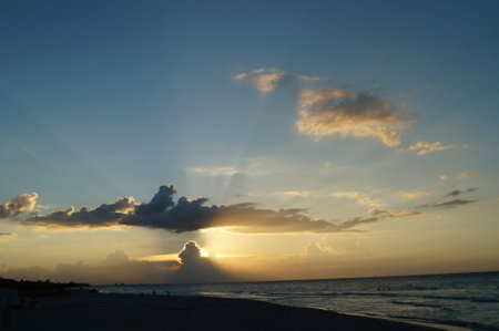 Golden sunlight spills across the horizon, casting a warm glow on gentle waves. Clouds weave intricate patterns against a backdrop of deep blue, creating a serene evening atmosphere.の写真素材