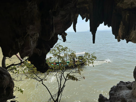 A small boat glides through calm waters just outside a jagged cave, framed by impressive rock formations and tropical greenery. The afternoon sun casts a serene light.の写真素材