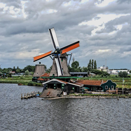 A striking windmill with orange and white sails towers over a serene river, surrounded by green...