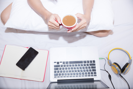Young woman drinking coffee at home in her bed and checking her laptop, top viewの写真素材