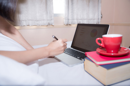 Happy casual beautiful woman working on a laptop sitting on the bed in the house.