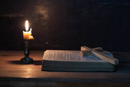 Wood cross laying on an opened Bible, with candle and candlelight on table