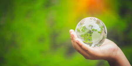 Human hands holding earth sphere crystal or sustainable globe glass with sunlight at green nature background in ecology environment forest. concept of conservation environmental, protection planet.の写真素材