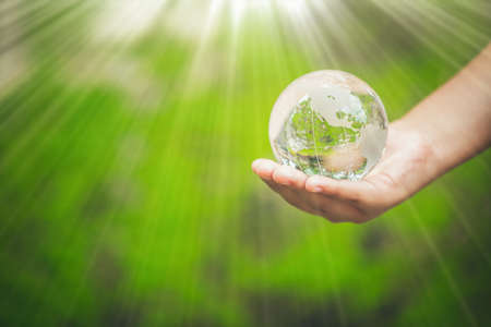 Human hands holding earth sphere crystal or sustainable globe glass with sunlight at green nature background in ecology environment forest. concept of conservation environmental, protection planet.の写真素材