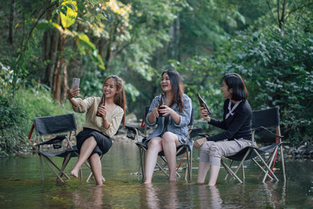 group of Asian girls enjoying a day at the  during holiday camping . Sitting on a camping chair Dip them legs in the River and drinking cold beer from the bottle . Vacations, holidays camping and relax concept.の写真素材