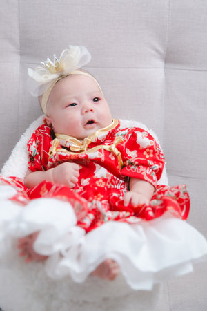 Closeup cute newborn baby in red bodysuit lying down alone on bed. Adorable infant rests on white bedsheets, staring at camera looking peaceful. Infancy, healthcare and paediatrics, babyhood concept.の写真素材