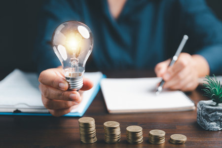 Businesswoman hand holding lightbulb with using calculator to calculate and money stack. Save energy and money with accounting finance in office concept. Idea of energy saving planning in home.の写真素材