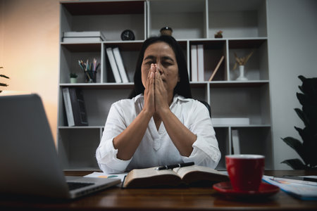 Businesswoman Praying with Eyes Closed. Businesswoman with her hands folded waiting for good news sitting at workplace at the table in the office. Hands folded in prayer gesture beg about something.の写真素材
