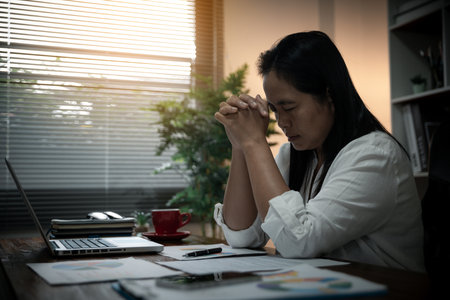 Businesswoman Praying with Eyes Closed. Businesswoman with her hands folded waiting for good news sitting at workplace at the table in the office. Hands folded in prayer gesture beg about something.の写真素材