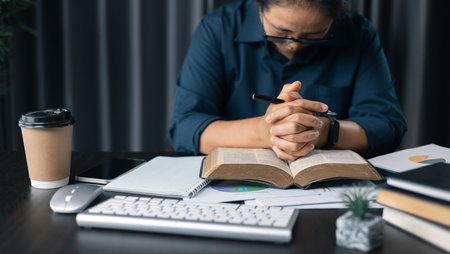 Woman with hands clasped praying while sitting at desk in office. Businesswoman praying at work. Contemplative prayer thinking in office. Hands folded in prayer gesture beg about something.の写真素材