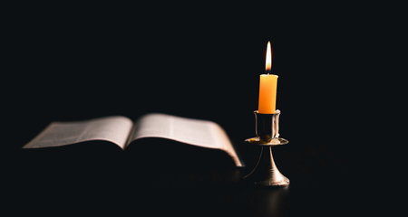 Dark, an antique candle cast its light on the old Bible, its flames flickering gently over the book on the table, symbolizing the enduring essence of religion. candle, bible, dark, light, book, old.の写真素材