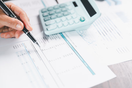 Auditor reviews financial statement and balance sheet on desk using a calculator, representing the...