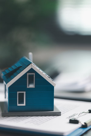 Real estate agent and customer are discussing contract to buy house, with house model in background symbolizing real estate, insurance, and loan arrangements involved in process.の写真素材