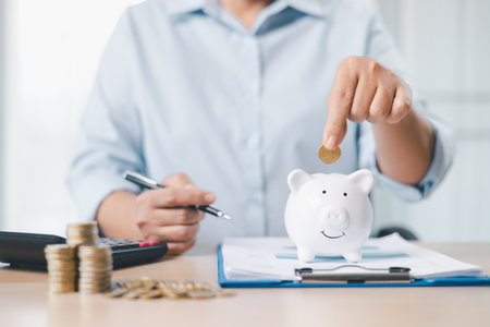 Concept of saving money and investment planning. A businesswoman drops coin into white piggy bank on office desk with documents, symbolizing financial security, budget management and future wealth.の写真素材