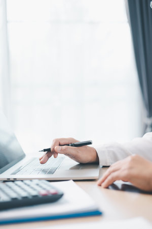 Businesswoman works on laptop while holding pen and reviewing financial documents at office desk with calculator. Concept of accounting, business planning, financial analysis and productivity.の写真素材