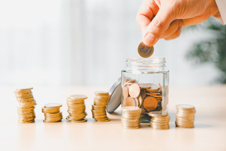 Hand places coin into saving jar, representing how smart finance, investment, banking practices turn money, cash, currency into financial wealth drives business growth and strengthens the economy.の写真素材