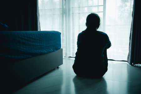 A lonely woman sits on the floor by window looking outside in dark room. Concept of depression,...