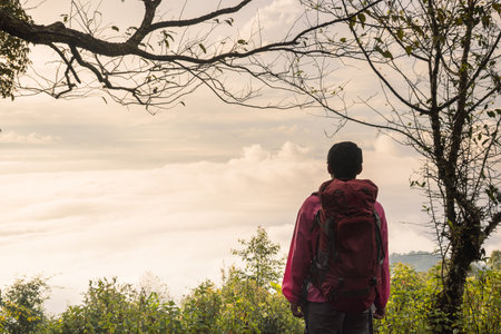 Traveler with backpack standing on mountain top looking at scenic sunrise over sea of mist,...