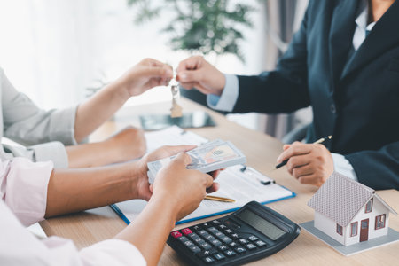 Close up of real estate agent handing house key to client with contract paperwork, calculator and...