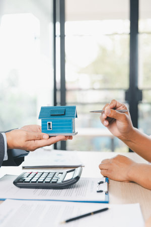 Close up of real estate agent showing small house model to client with calculator, contract paperwork on desk, concept of mortgage loan, home ownership, property investment agreement. mortgage loanの写真素材