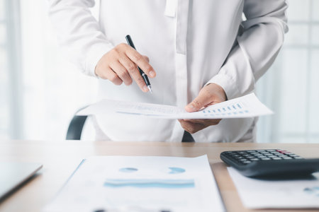 A businesswoman reviews financial report while holding documents and pointing with pen at office desk. Concept of accounting, budget management, financial analysis and corporate business planning.の写真素材