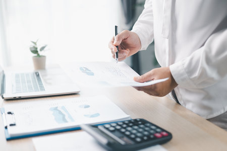 A businesswoman reviews financial report while holding documents and pointing with pen at office desk. Concept of accounting, budget management, financial analysis and corporate business planning.の写真素材