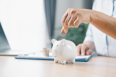Concept of saving money and investment planning. A businesswoman drops coin into white piggy bank on office desk with documents, symbolizing financial security, budget management and future wealth.の写真素材