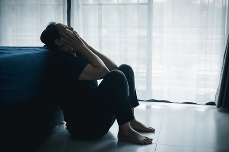 A lonely woman sits on the floor by window with hand on head in dark room. Concept of depression, sadness, anxiety, stress, mental health problem, loneliness and emotional struggle in life.の写真素材