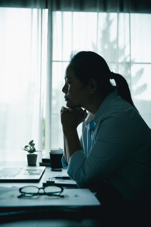 Stressed businesswoman working late at home office, feeling exhausted while reviewing financial documents on laptop. Concept of work stress, burnout, deadline, and overwork.の写真素材