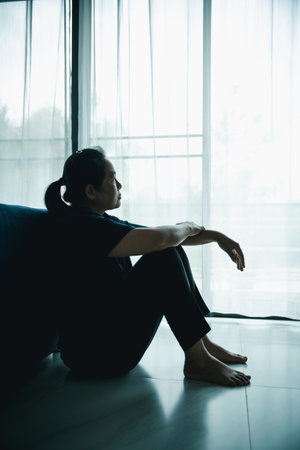 A lonely woman sits on the floor by window looking outside in dark room. Concept of depression, anxiety, sadness, loneliness, mental health problem, emotional struggle and hope for better life.の写真素材