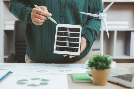 Eco-friendly office scene with person working on laptop near solar panel and wind turbine model, representing green business, renewable energy innovation and sustainable technology workspace.の写真素材