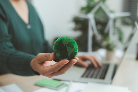 Eco-friendly office scene with person working on laptop near solar panel and wind turbine model, representing green business, renewable energy innovation and sustainable technology workspace.の写真素材