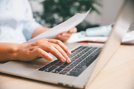 Office worker typing on laptop while reviewing documents, illustrating digital workflow, business tasks, online work, productivity and professional administration in modern workplace environment.の写真素材
