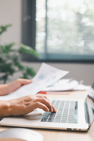 Office worker typing on laptop while reviewing documents, illustrating digital workflow, business...