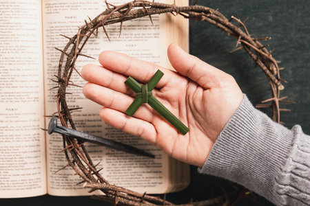 Hand holding palm leaf cross above open Bible, crown of thorns, iron nail, symbolizing Christian faith, Palm Sunday, sacrifice, Easter reflection, religious background for worship, prayer concepts.の写真素材