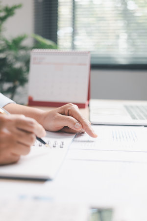 Office worker analyzing charts while pointing at data, writing notes in notebook, illustrating review, planning, strategy, productivity, professional workflow in modern workplace.の写真素材